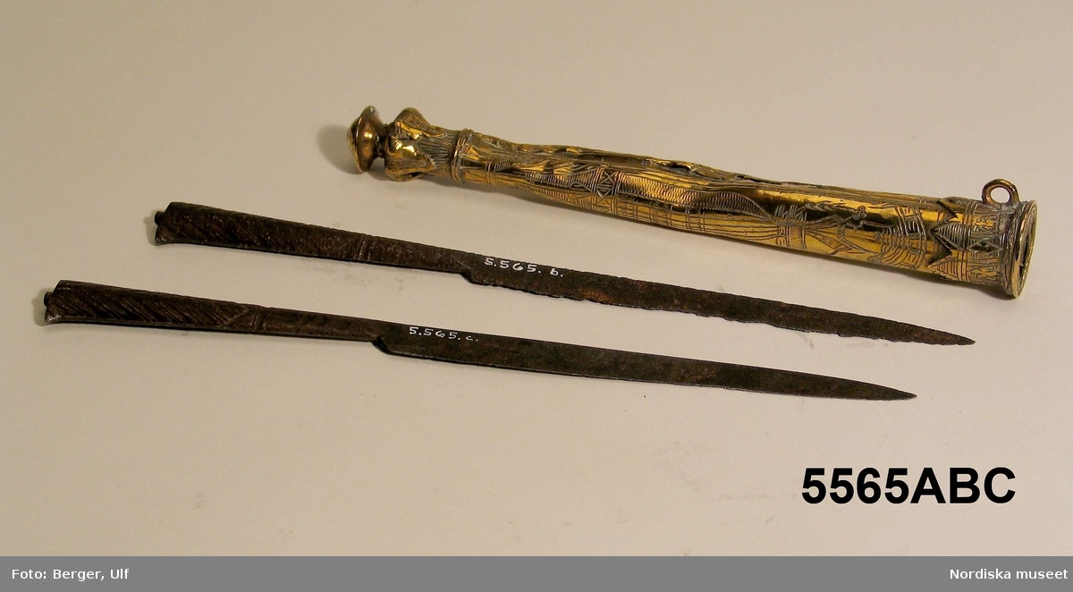 Montertext i Dukade bord:
Knivpar av järn i slida av graverad mässing. 1500-tal. "Kvinnobestick" som bars i en lång kedja eller rem hängande från bältet. 
a) Knivslida
b) kniv
c) kniv