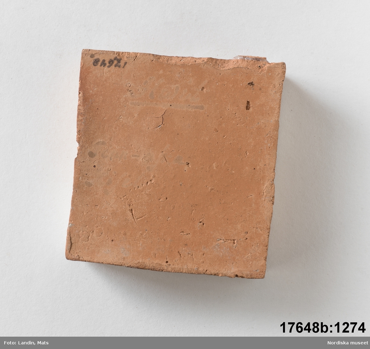 Kvadratisk platta av bränd röd lera, ena sidan glaserad i vitt. den andra sidan oglaserad. På den oglaserade sidan text (svagt och därför svårtydd): Tenn-aska / Kisel" [?] . 
/Leif Wallin 2014-01-07