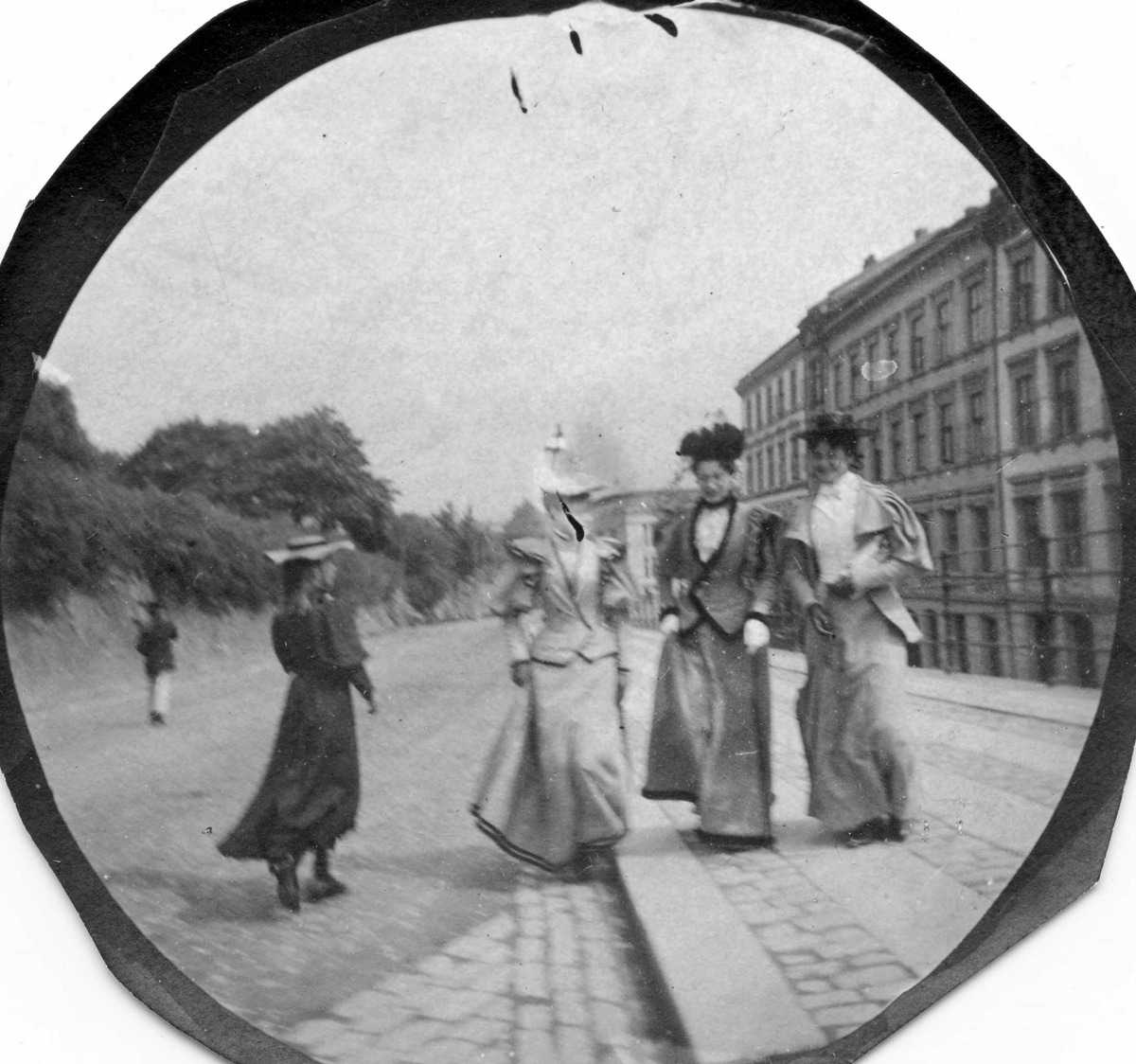 Fire unge kvinner spaserer i bygate, Oslo, tre vendt mot fotografen. Bygårder på den ene siden av gaten bak smijernsgjerde, park på den andre. 