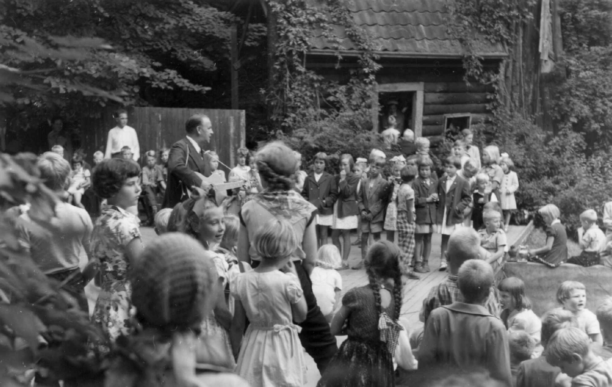 Alf Prøysen med gitar spiller og synger på friluftsscenen på Norsk Folkemuseum i 1954. Stort publikum med barn og voksne.