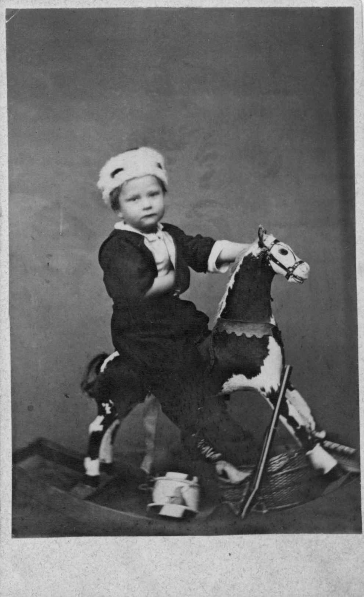 Portrett av gutt på gyngehest. Gutten har en lue på hodet. Uten oppl.
Fra album som har tilhørt fotograf Chr. Wær(e)nskjold eller hans svoger.