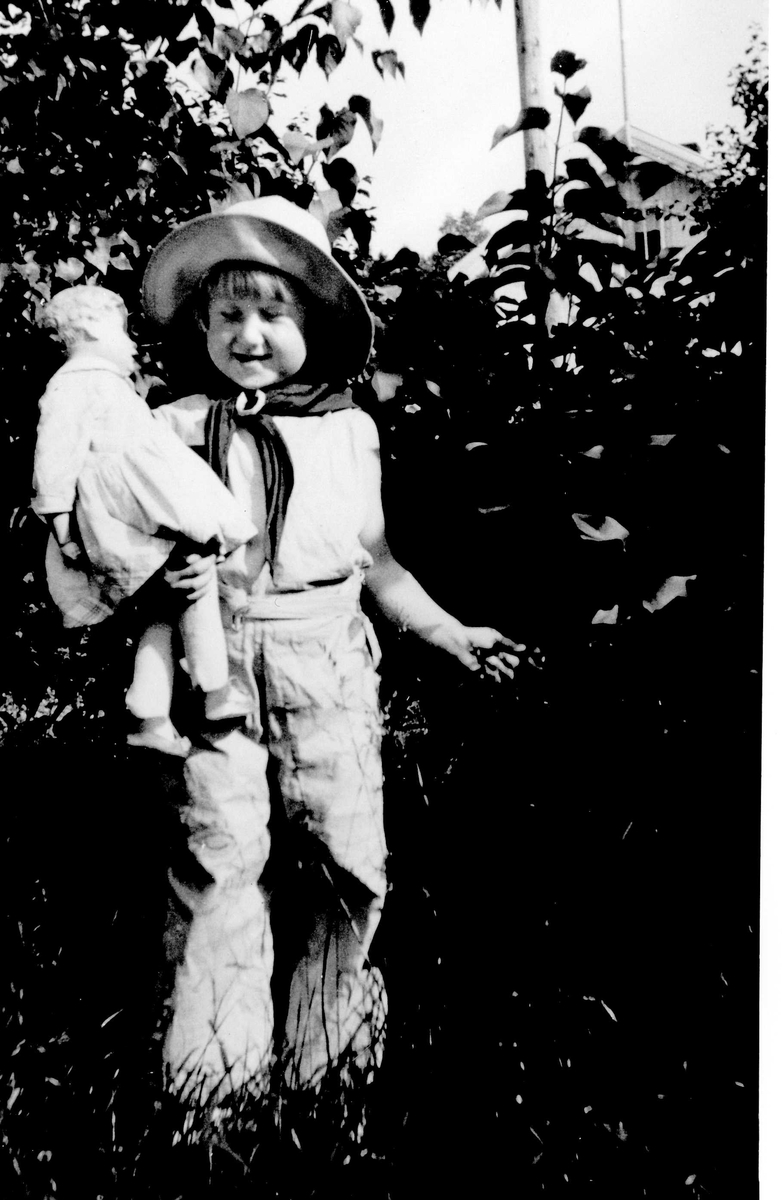 Pike med dukke i hagen. Jorunn Fossberg i sommerhusets hage på Nærsnes, Røyken, Buskerud ca.1932. Hun er ikledd moderne strandpysjamas i rosa pikestoff og solhatt kjøpt av far i Paris.
Fra Jorunn Fossbergs private familiealbum.