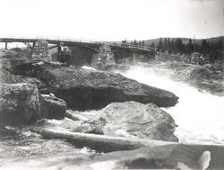 To mennesker står foran bro med steinpilarer ved foss.