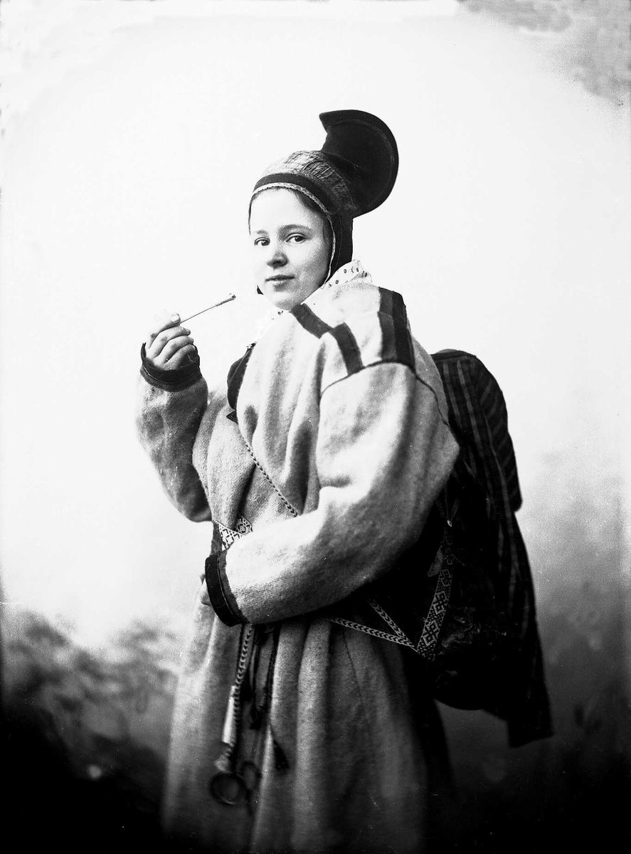 Kvinne ikledd samiske tradisjonelle klær: kofte, hornlue og komse på ryggen. Pipe og beltering med nålehus og fingerbøl.
