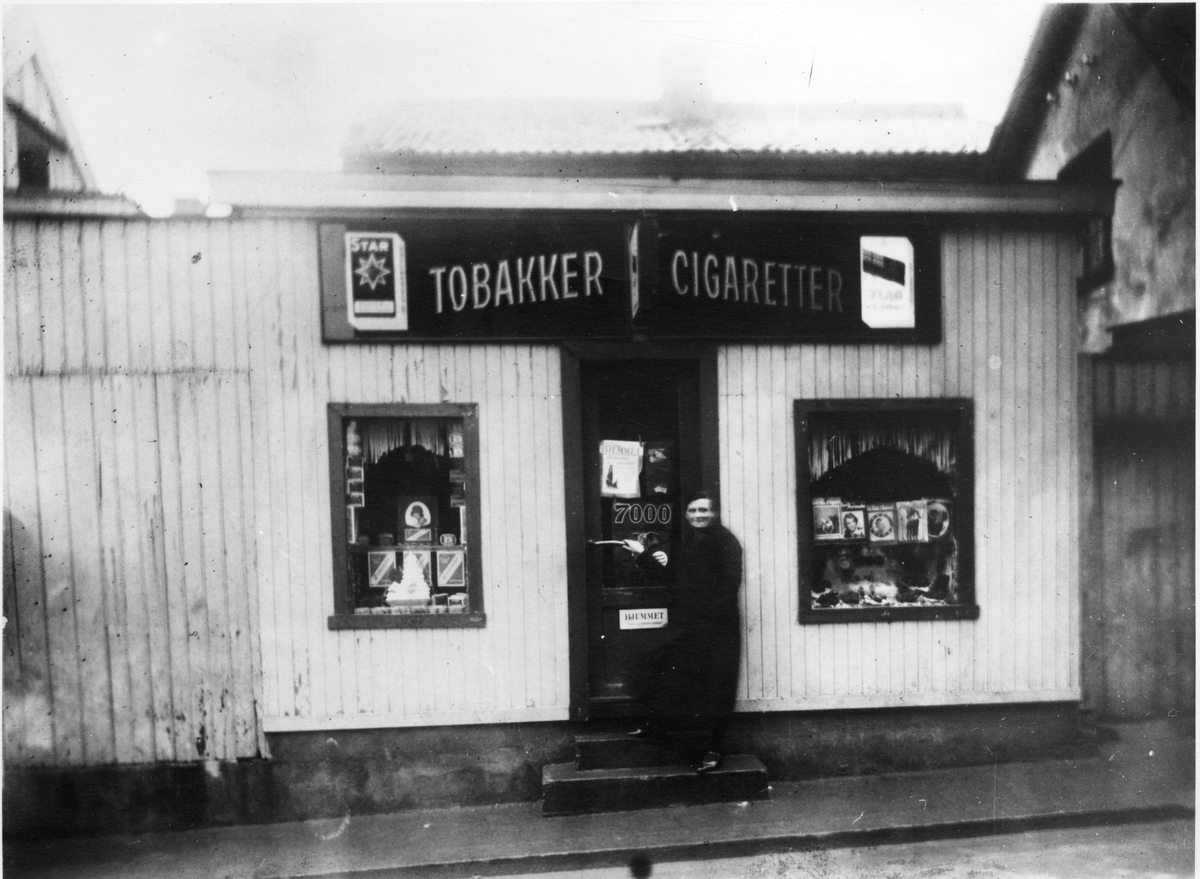 Gatebilde. Kvinne ved inngangen til kiosk. Skilt med reklame for tobakk og sigaretter.