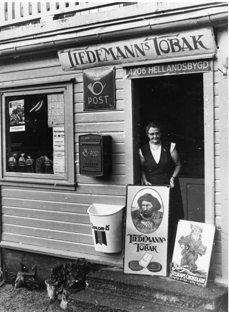 Kolonial og post i Hellandsbygd. Kvinner viser frem et skilt med reklame for Tiedemanns Tobak.