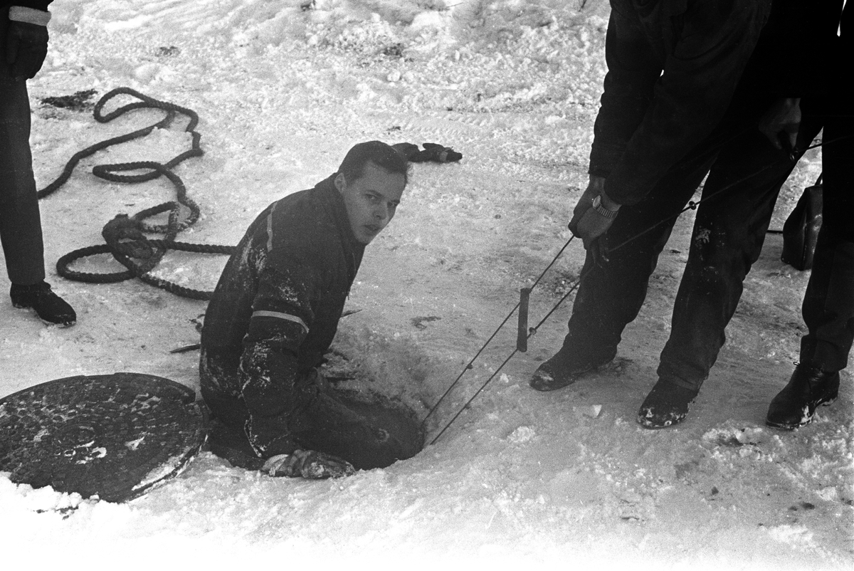 Redningsmann på vei opp av en kloakk-kum etter å ha reddet en hund da den falt i kloakken på Sjølyst i Oslo i januar 1961.