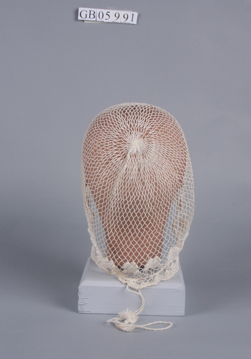 Trukket sammen i kronen. Broderte blomster langs kanten. Kant av tettere masker. Trekkebånd med dusker.