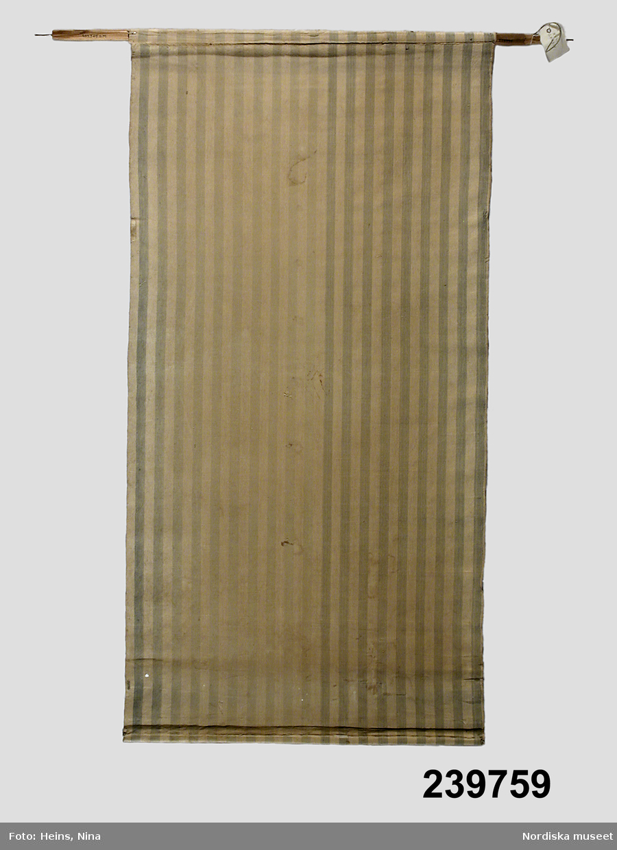 Vertikalrandigt bomullstyg i blått och vitt. Trästång med spikar i ändarna, nedre käpp (rundstav) i sydd kanal. L. 111, 5 cm., B. 84/56 cm.
Anm.: Fläckar, blekt, gammal lagning.
/Marie Jansson-Lohse 2006-10-02