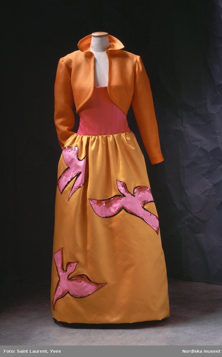 Hellång aftonklänning med axelbandslöst liv av sidenduchesse. Livet ceriserosa, kjolen orangegul med applikationer i form av stiliserade fåglar i rosa siden prydda med paljetter och pärlbroderi. Från Yves Saint Laurents vårkollektion 1988. Nordiska museet inv nr 318887.