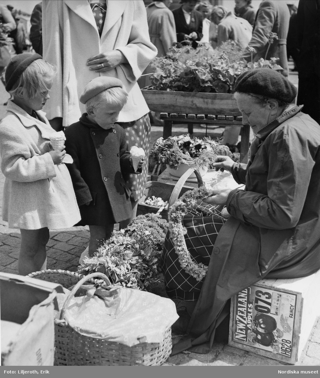 Två glassätande barn som tittar på en äldre kvinna som säljer blommor
