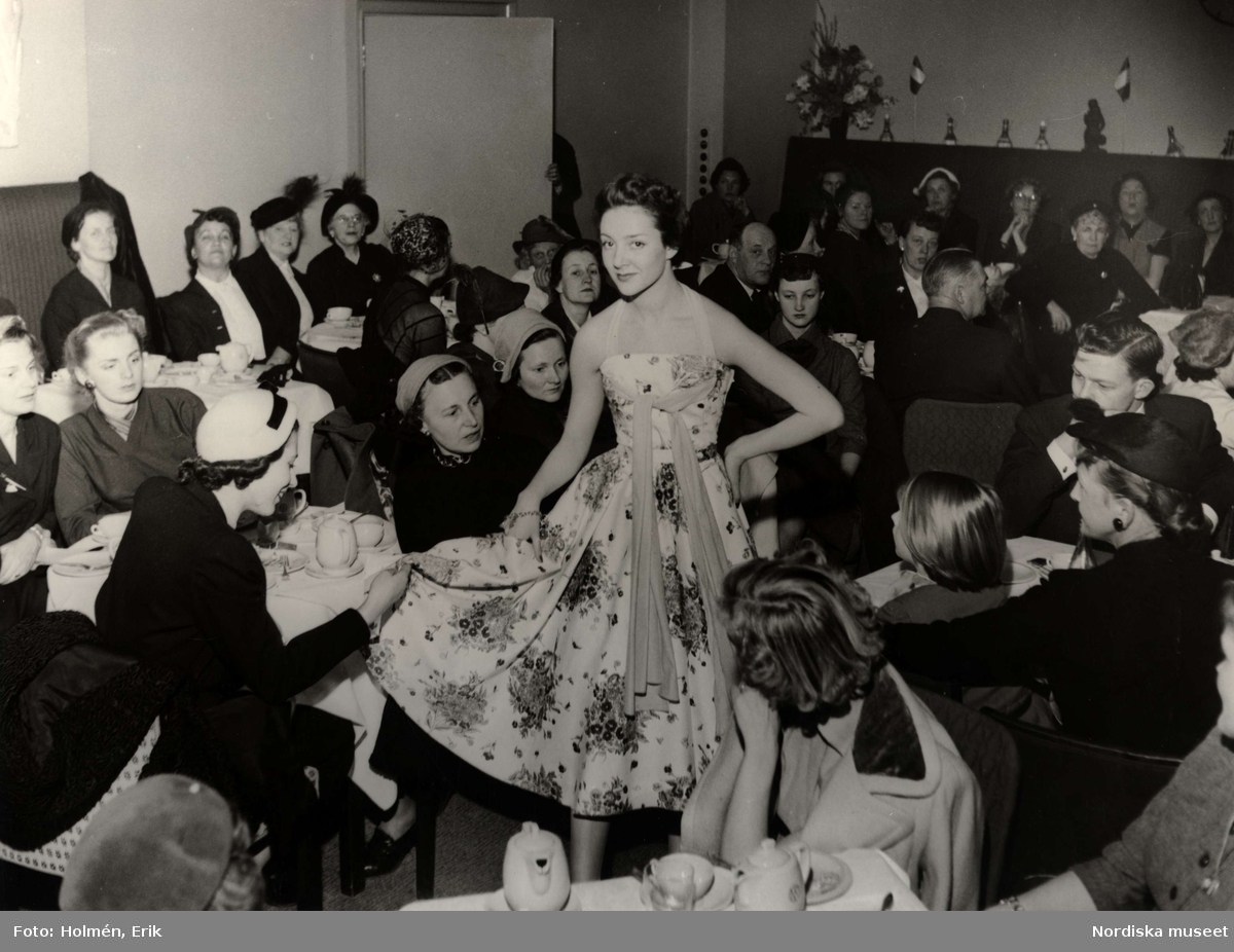 Visning Italiana på NK (Nordiska Kompaniet) 1953. Kvinnlig modell iklädd ljus klänning med bara axlar och blommönster. Modellen står i rum med publik som sitter vi små bord dukade med kaffe- eller tekoppar och kannor.