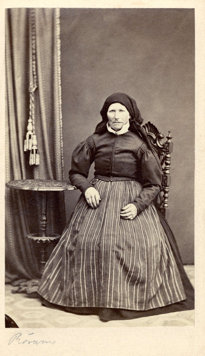 En äldre kvinna i schalett sitter på en stol och vilar armen på ett litet pelarbord. Rörum. Nordiska museet inv.nr. 18533F.
