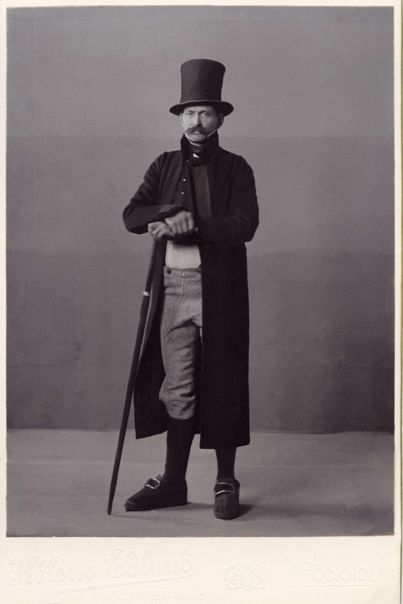 En man poserar i folkdräkt från Fellingsbro socken med lång rock och hög hatt.