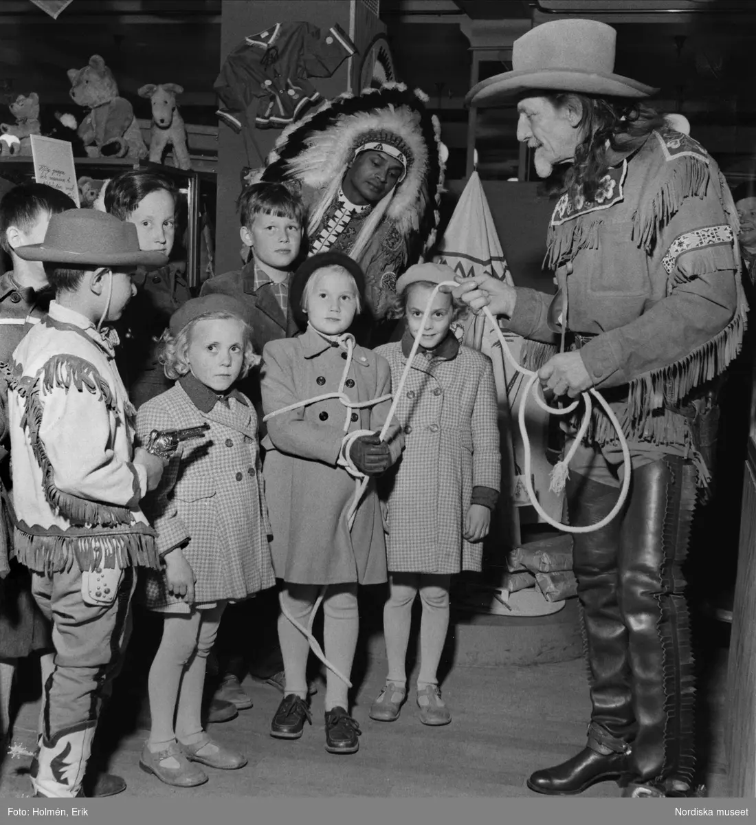 "Buffalo Bill på leksaksavdelningenl".  Cowboy och indian tillsammans med barn på Nordiska Kompaniets leksaksavdelning 1953.