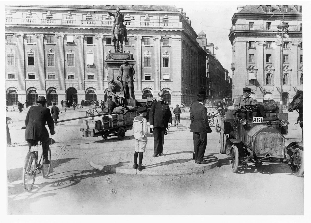 Stadsbild från Gustav Adolfs torg i Stockholm, 1910. Mitt i rondellen står en polis som övervakar trafiken, några kostymklädda herrar och en pojke i sjämanskostym med korta byxor. Cyklister, en bil och hästdragna kärror samsas på gatan. I bakgrunden byggnader och L'Archevêques staty förställande Gustav II Adolf.