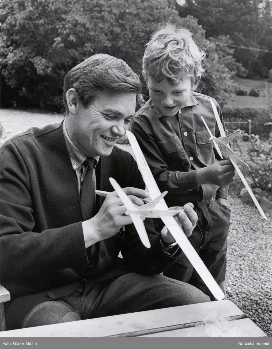 Porträtt av skådespelaren Lars Ekborg (1926-1969) tillsammans med sonen Dan Ekborg (f. 1955). Lars Ekborg sitter vid ett bord utomhus, i händerna håller han ett modellflygplan, sonen står vid hans sida, även han håller i ett flygplan. De ler och samtalar med varandra.