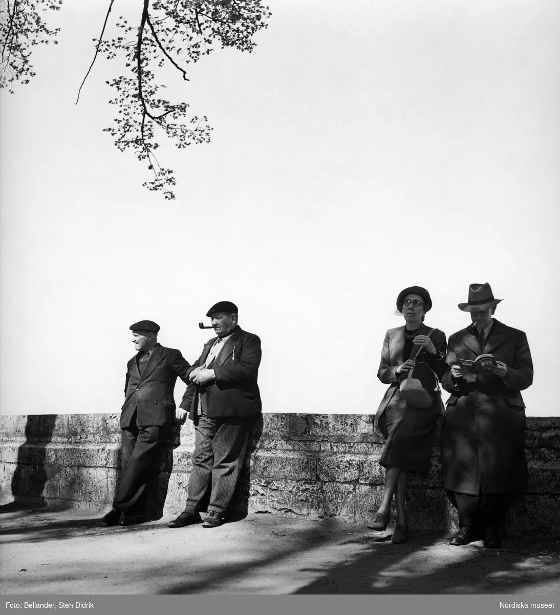 Två äldre män med basker på huvudet och ett par i övre medelåldern står lutade mot en stenmur.
Mannen har en cigarett i munnen och bläddrar i en guidebok om Chartres.