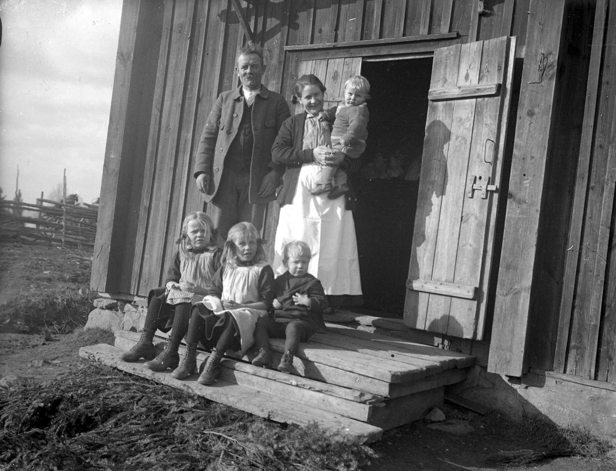 Gruppbild av en familj på trappan till sitt hus. Tre barn sitter, mannen och kvinnan står. Kvinnan håller ett litet barn på armen. I bakgrunden skymtar en gärsgård.