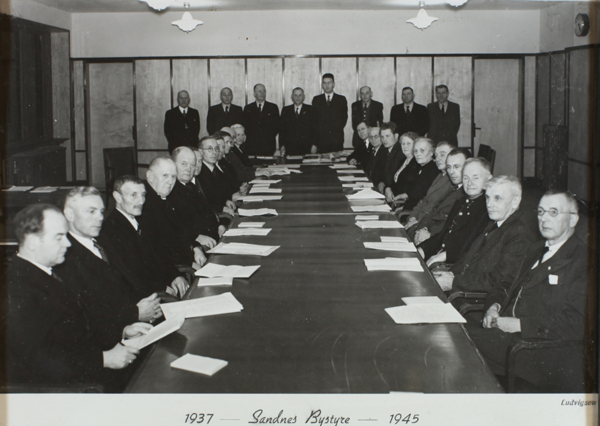 Sandnes Bystyre 1937-1945: menn og kvinner sittende ved et langbord, papirer liggende på bordet, åtte menn stående foran bakveggen.