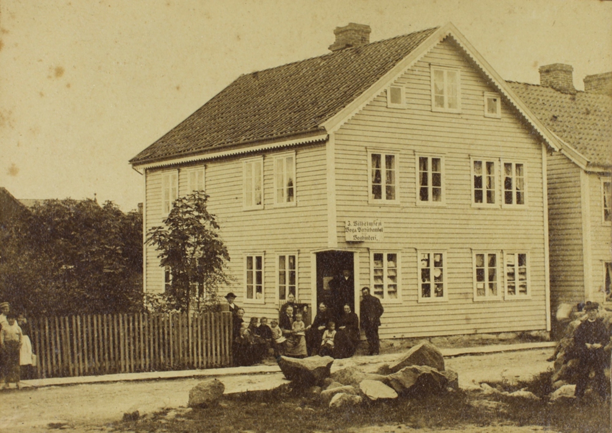 Familie foran et bolighus (J. Wilhelmsen Bog & Papirhandel. Bogtrykkeri).