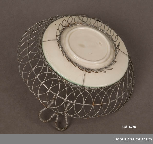 Assiett av keramik (majolika) med kantgaller i järntråd, något rostat.
Jämför UM18237.