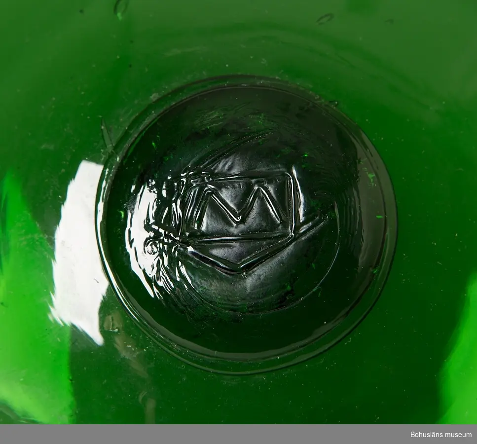 Sex gröna glasflöten.
Försett med isatt märke med bomärkesliknande emblem. "M" inom rektangel.