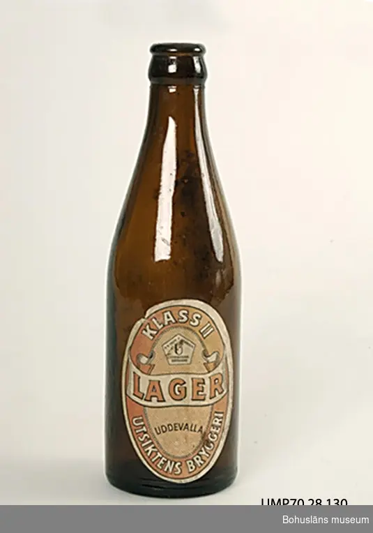 Flaska av brunt glas med alkamynningsknopp,  det ringformade  urtaget på flaskknoppens utsida för "Alkaförslutning" i aluminium. 
Innehåll 1/3 liter, en s.k. "kvarter".
Oval pappersetikett,  8,8 x 6,2 cm, med röd och svart text på vit botten:
Klass II 
Lager 
Utsiktens bryggeri
Uddevalla. 

Alka-förslutningen är ett svenskt patent av A. J. Jonsson, Linköping introducerad efter 1930.
Ölflaskan är Sveriges andra standardiserade bruksföremål, 
d.v.s ett föremål med i landet överenskomna mått oavsett tillverkare.  Sveriges första "standard" är tändsticksasken.

Från Internetkälan Uddevallabloggen är historiken om Utsiktens bryggeri hämtad, skriven av Gunnar Klasson, Uddevalla:
"Idag [år 2007] finns det inga bryggerier i Uddevalla men annat var det förr. Ett av de äldsta var Utsiktens bryggeri från 1854 tätt följt av Svante Natt och Dags bryggeri i kvarteret Dagson. Natt och Dags bryggeri var länge det största som moderniserades och byggdes ut 1902. Men 1924 var konkursen ett faktum och bryggeriet lades ned. Samma år köpte Schwartzman & Nordström AB bryggeribyggnaderna och byggde om dem till konfektionsfabriken.
Utsiktens bryggeri låg mellan Underåsgatan och Vintergatan på Väster. Bryggeriet fick sitt namn efter platsen som sedan länge kallades för Utsikten. På den tiden fanns det ingen bebyggelse utan då man stod på höjderna vid Annegreteberg (nuvarande Uddevalla sjukhusområde) hade man en strålande utsikt ut mot Byfjorden.
På platsen låg i vart fall sedan 1830-talet ett brännvinsbränneri. Innehavare var den kände 
Uddevallabon C A Kullgren, han med stenindustrin. 
Kullgren avled på världsutställningen i London 1851 och hela bränneribranschen var vid den här tiden i gungning. De flesta småbrännerierna runt om i Sverige lades ned och produktionen koncentrerades till större enheter.
Utsiktens brännvinsbränneri konverterades därför 1854 till ett ölbryggeri. På en del håll sågs detta till och med som en nykterhetsbefrämjande åtgärd.
I början hette det Uddevalla Ångbryggeri AB men namnet ändrades sedan till Utsiktens 
bryggeri. 1876 brann bryggeriet ned till grunden. Innehavaren hette då Kjellman. Det var 
denne Kjellman som byggde den bryggeribyggnad som gamla Uddevallabor minns.
Bryggeriet bytte då och då ägare. Kjellman sålde bryggeriet till Konrad Forster och senare 
ägare var J L Larsson, Arvid Uddenberg och Nils Sjunneson. 
1919 köpte AB Pripp & Lyckholm Utsiktens bryggeri och de nya ägarna genomförde 1926 en omfattande modernisering av anläggningen. Den 20 september visades det nya bryggeriet för särskilt inbjudna och för allmänheten dagen därpå. 
Av en broschyr från det tillfället framgår det vilka produkter man tillverkade. Det var Pilsner, Extra lager och s k Skattefritt (lättöl?). Man hade också en omfattande tillverkning av läskedrycker som Vichyvatten, Seltersvatten, Sockerdricka, Lemonad, Hallonlemonad, Champagnedricka (Champis?), Pommac, Messina, Monark, Materna, Stadion, Pommona, Citro, Banancider, Fruktcider, "Bock-öl" och "MÃ¼nchner-öl".
Mitt i smällkalla vintern, den 27 januari 1942, inträffade en storbrand i bryggeriet. Brandmännen kämpade i 20 graders kyla och gjorde ett gott arbete, men trots det fick bryggeriet omfattande skador.
Uddevallas mångårige brandchef Eric Ström berättade om branden vid en stadsvandring i  kvarteren på Väster ---. "Plötsligt var han ensam på  platsen! Var var brandsoldaterna? Nåväl det värsta var över och efter en stunds letande fann han dem nere i bryggeriets lager där de värmde sig både invändigt och utvändigt. Samtliga  hade var sin "stor stark" som de halsade då han dök upp".
Efter branden började man omgående att reparera byggnaden och man passade på att förnya maskinparken samtidigt.
Pripp & Lyckholm lade ned produktionen vid bryggeriet 1961 och under några år fungerade 
lokalerna som nederlag och distributionscentral. Det var då fortfarande liv och rörelse på  platsen men 1968 hade Pripps byggt ett nytt lager på Kuröd dit man flyttade verksamheten. 
Bryggeribyggnaden stod kvar flera år men jag hittar ingen uppgift om när den revs." 

Litt: Fogelberg, T., Nisbeth Å.: Värmländska glasbruk. 2, Liljedals glasbruk /
Värmlands museum 1979. Småskrifter utgivna av Värmlands museum.
Förpackningar. Kulturen 1987, Årsbok för Kulturen i Lund.
Glasfolket. Buteljglasbruket i Hammar 1854-1930 . Örebro, Morgonstjärnan. Del I 1991, Del II 1994.
Hermelin, C. F., Welander, E.: Glasboken. Historia, teknik och form. Askild & Kärnekull 1980.
Internetkälla: Uddevallabloggen.
