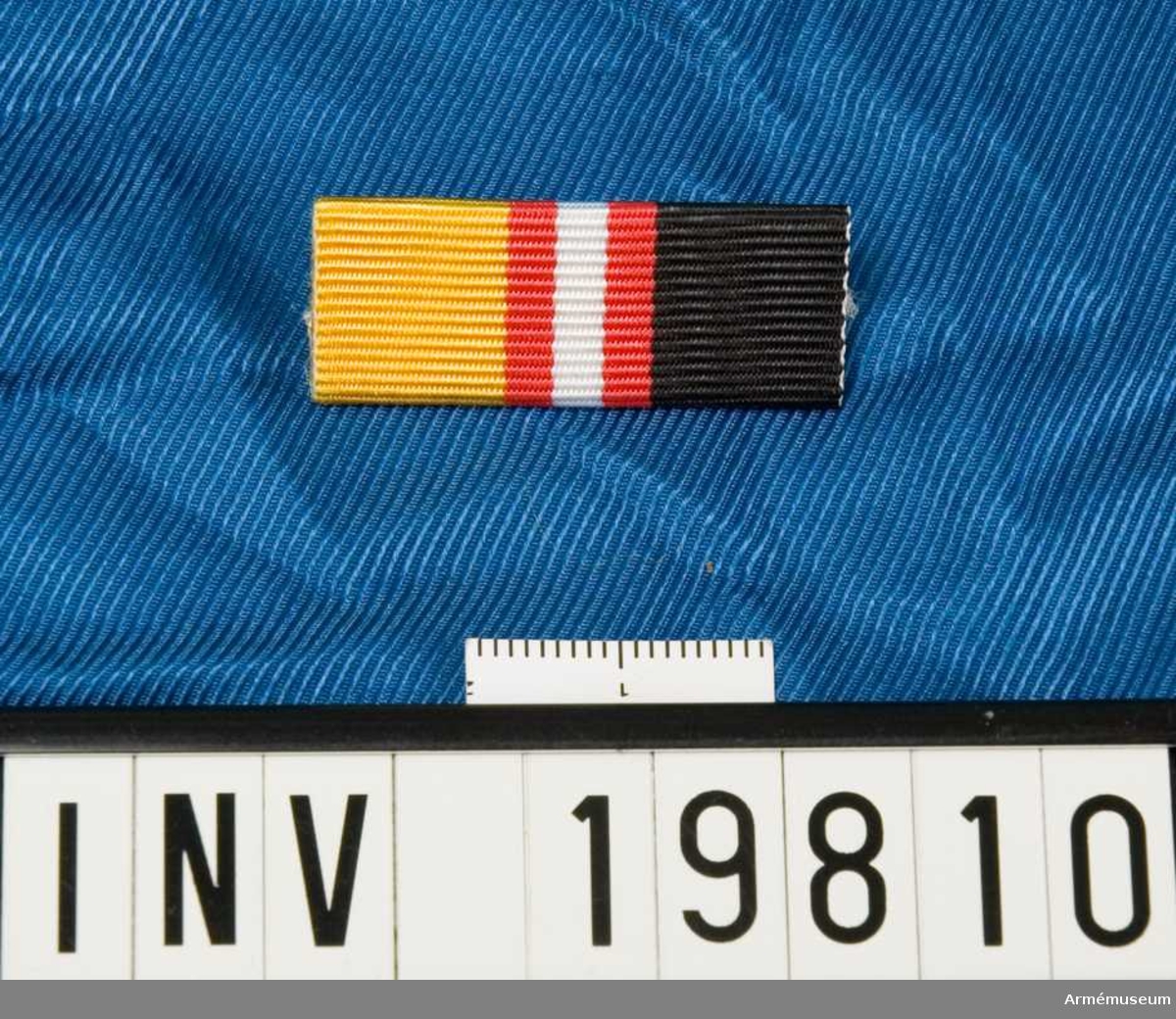 Band kluvet i svart och gult med en vit rand i mitten åtföljd av en röd rand på vardera sidan. Släpspännet förvaras i ask tillsammans med en medalj och en miniatyrmedalj.