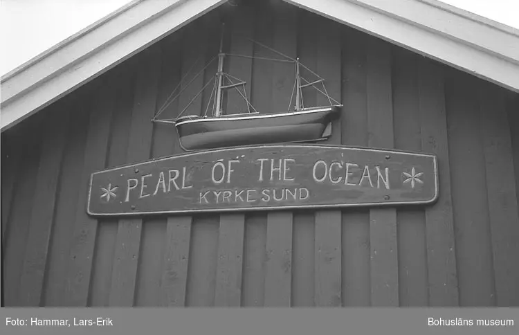 Namnbräda "Pearl of the ocean Kyrkesund" och modell av fiskebåt på utsidan av en fiskebod i oktober 1978
