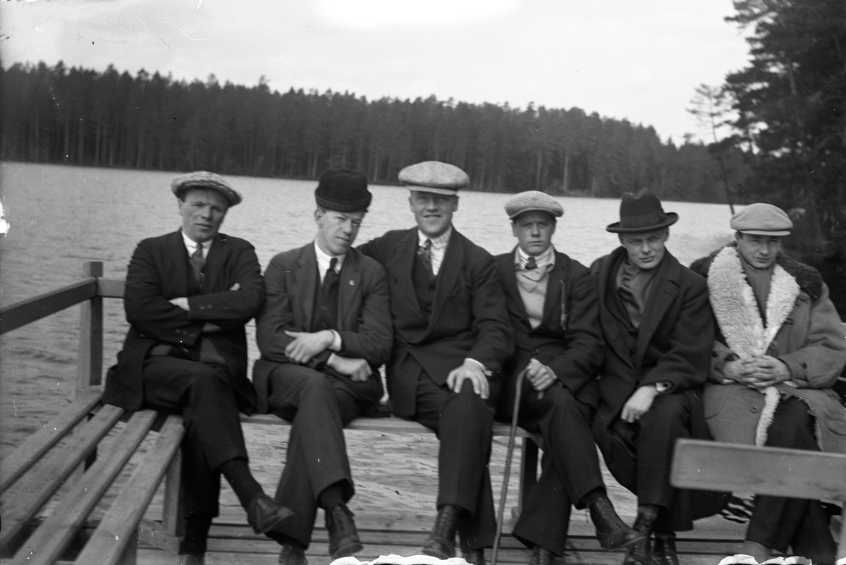 Lungkliniken i Eksjö. Sex män, Gustav Andersson (6), sitter på en bänk på en brygga vid sjön. Barrskogen skyns i bakgrunden.