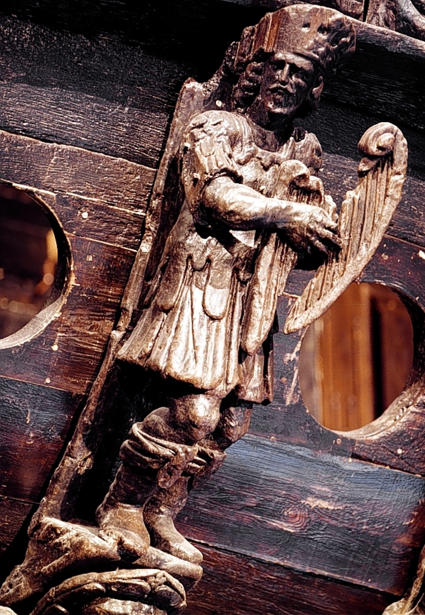 Skulptur utformad som en harpspelande man, med skägg och långt hår. Skägget är svagt krusigt och håret faller i lockar ned mot axlarna. Huvudet är vridet mot höger. Mannen står i knäande ställning, med höger ben något framskjutet och är iförd romersk rustning av typ muskelpansar - Thorax - med nedhängande lambrequinliknande och läderimiterande flikar ovanpå den tjocka livklädnaden eller tunican.

Figuren är iförd en stövelliknande fotbeklädnad med tjocka skaftprydnader. På huvudet förefaller han bära en slags mössa kring vilken en öppen krona trätts. I händerna, som hålls lyfta framför bröstet, håller han ett litet trekantigt stränginstrument, liknande en liten harpa och prytt med ett svanhuvud.

Mannen står på en bastant sockel med trekantig profil. Sockelns sidor är ornerade med enkla ritsar. Figuren är skuren i ett med sockeln och liksom det profilsvängda, smala bakstycket, snidad i mycket hög relief, på gränsen till rundskulptur. Bakstyckets under- eller baksida, som alltså blir skulpturens baksida, är slät och obetydligt snedskuren i sidled.

Skulpturen är förhållandevis välbevarad. Förutom allmän nötning är det endast smärre partier, huvudbonaden, händerna och bakstyckets överdel som är kraftigare skadat.

Text in English: Sculpture of a man with beard and long hair, dressed in Roman armour, playing a harp.
The man''s head is twisted to the right and he stands with his knees slightly bent, with the leg somewhat obtruding. He is clad in Roman armour of the thorax type, with hanging lambrequin-like simulated leather flaps outside his tunic. He is wearing boot-like footgear with thick, decorated tops.

On his head he appears to be wearing a kind of hat or helmet, round which there is an open crown. The beard is lightly curled and the hair falls in curls over his shoulders. His arms are held across his chest and in his hands he is holding a small, triangular, stringed instrument, similar to a small harp, decorated with a swan''s head.
The figure stands on a robust base with a triangular profile and ornamented with simple cut tracery. The figure and base are carved from a single piece of wood along with the ''back-board''.

The reverse side of the sculpture is smooth and is curved or concave from the foot up to the neck of the man. Here the back section angles up and continues above to cover the top of the man''s head.

The sculpture is relatively well preserved. In addition to the general wear there are only a few parts of it, such as the headgear, hands and upper section of the ''back-board'', that are badly damaged.