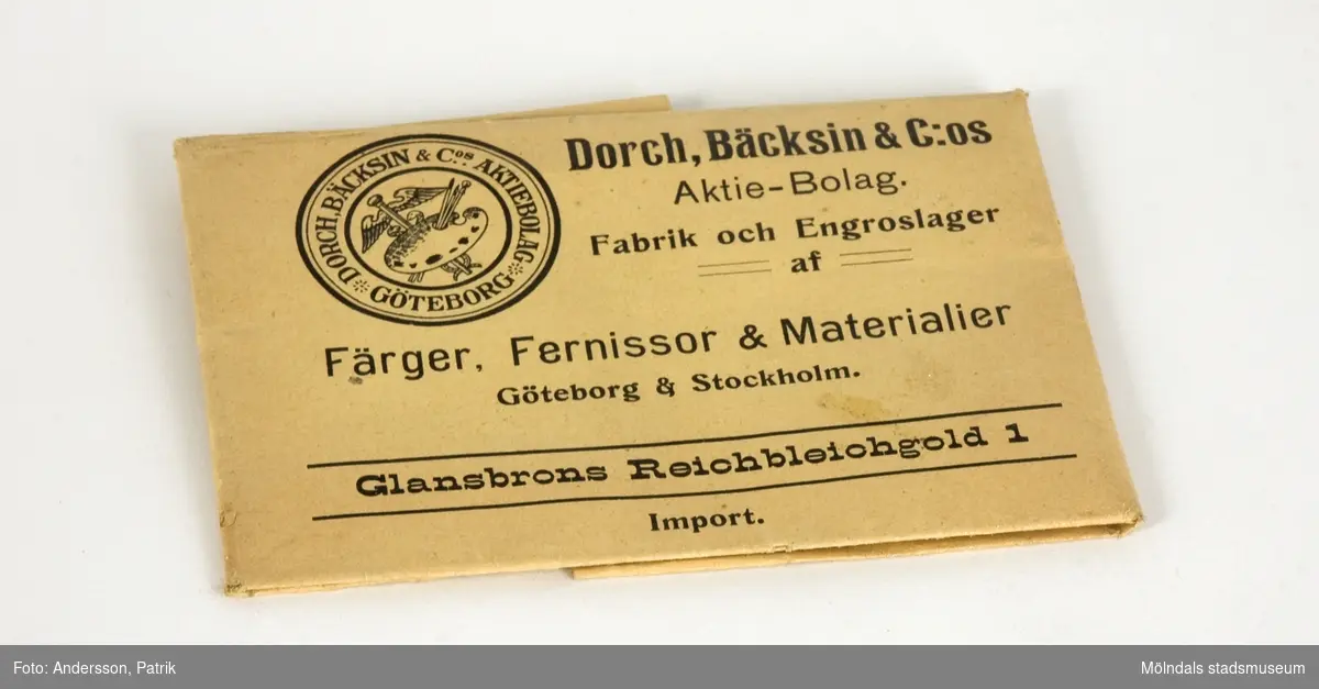 Förgyllningspulver, använt på Papyrus. Från ca 1910.Pulvret är invikt i två sorters papper. Innerst, ett vitt, glättat papper, inslaget i ett brunt papper med texten "Glansbrons Reichbleichgold 1", från "Dorch, Bäcksin & C:os Aktie-Bolag. Fabrik och Engroslager af Färger, Fernissor & Materialer, Göteborg & Stockholm". 