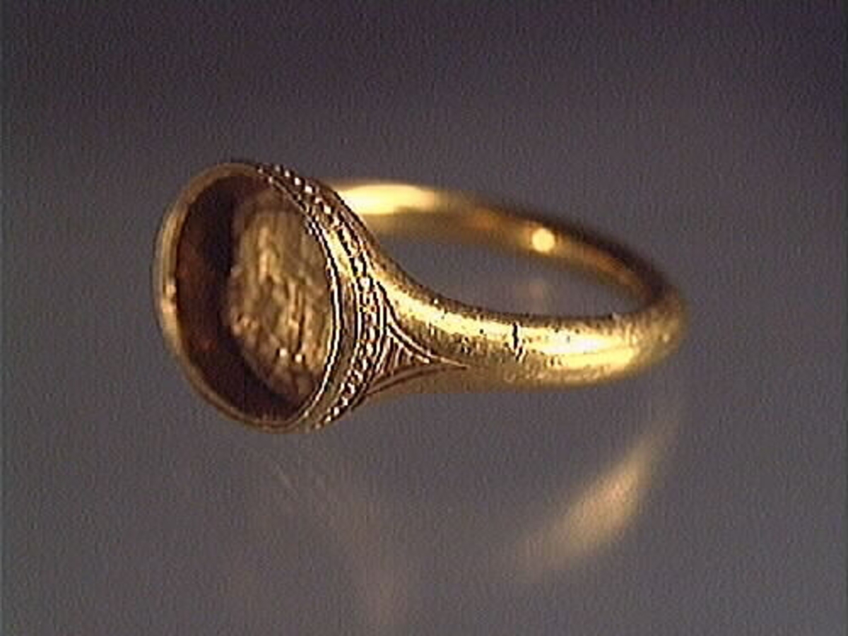 En fingerring av guld.
Guldringen har en oval infattning. Runt infattningen löper en pärlliknande bård. Bården består av 60 små och pärlliknande guldkulor, om 1 mm i diameter. På var sida om infattningen sitter en ingraverad, avsmalnande triangel försedd med horisontella streck. Ringen har varit försedd med en sten eller sigill vilken nu saknas. Ringens vikt uppgår till 10 gram.
Ringen är i mycket gott skick.

Text in English: Robust finger ring of gold with oval setting. The stone is missing.

Ingraved round the setting is a pearl-like border consisting of 60 tiny circles, 1 mm in diameter, and on either side of the setting - on the ring''s ''shoulders'' - is an inward curved triangle with horizontal lines.

Weight: 10 gram.
In very good condition.