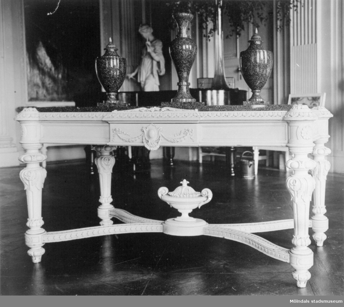 Ett ljust, snidat bord med tre urnor stående ovanpå. I bakgrunden ser man statyn "Våren" (i nordost) av Gioacchino Frulli (1766-1801). Stora salen på Gunnebo slott.