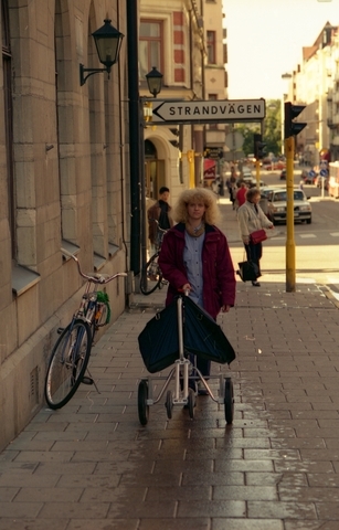 Brevbärare på Linnégatan i Stockholm. Brevbäraren bär vinröd jacka och blå byxor. Hon drar den större typen av postkärra med två stora och två små hjul. Kärran är även försedd med handbroms.