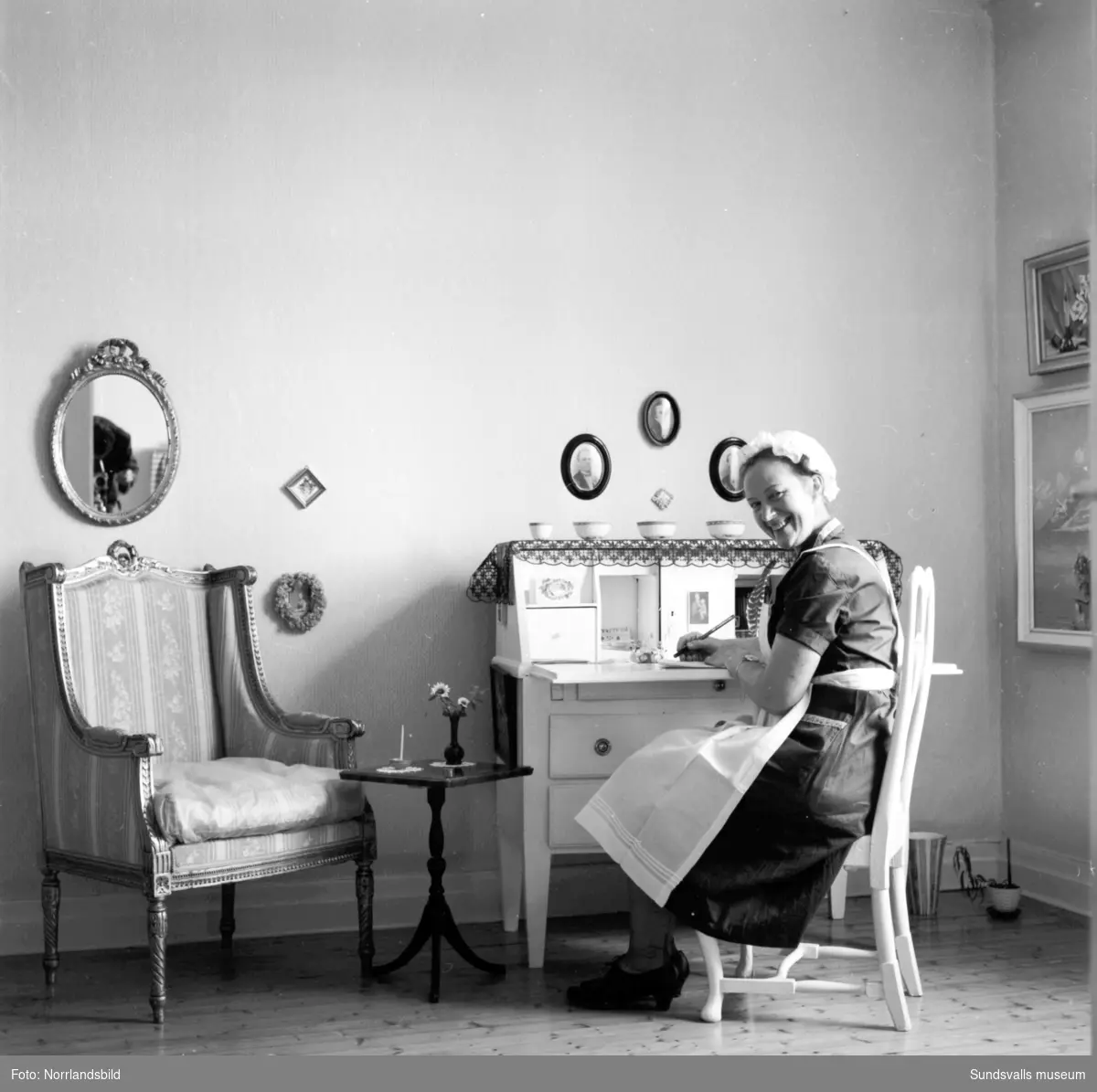 Kvinna i vitt förkläde och hätta fotograferad i köket och salongen. Svartvik.