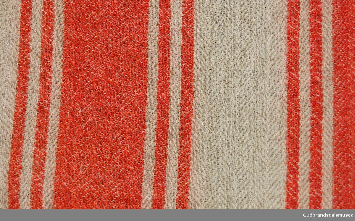 Rektangulært stykke vevd ull med striper i rødt på beige bakgrunn. Innlagt og sydd i kortendene. 