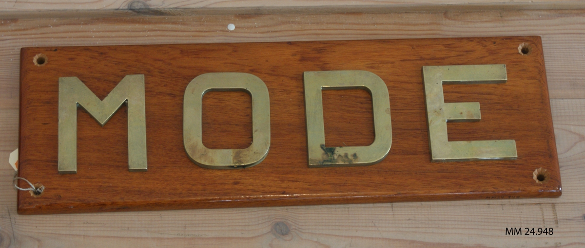 Fernissad träplatta. På framsidan bokstäverna MODE i guld. Alla hörnen genomborrade för upphängning.