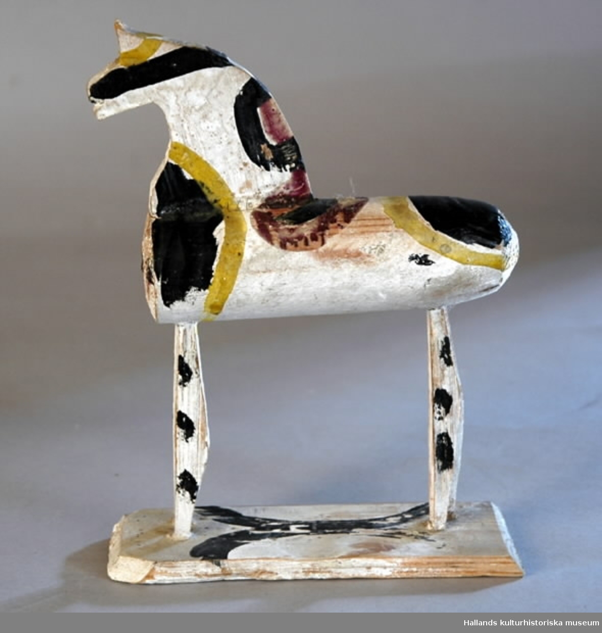 Leksakshäst av trä, vitmålad med mönster i gult, svart och gredelint. Kroppen cylindrisk, svansen har fallit bort. Huvudet och benen är profiler utskurna ur en träskiva. Hästen står på en rektangulär platta.