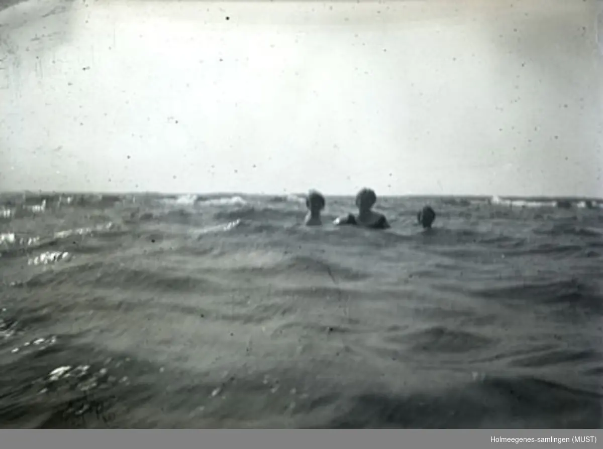 Tre personer bader i sjø med små bølger (bare hode og skuldre stikker opp av vannet). Motlys på bildet gjør det vanskelig å identifisere personene. Fint vær med lette skyer på himmelen. Muligens samme sted og tidspunkt som ST.K.HE 2007-011-0057 og -0058. Motivet er fremkalt på postkortpapir.
