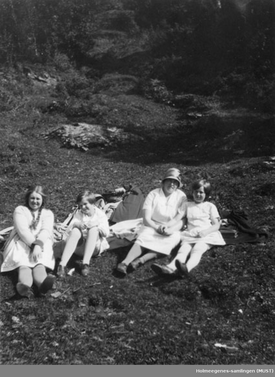 En kvinne og tre jenter i sommerkjoler sitter på pledd el.l. på bakken utendørs. Kvinnen har bøttehatt på seg. Se også ST.K.HE 2007-011-0111 til -0126.