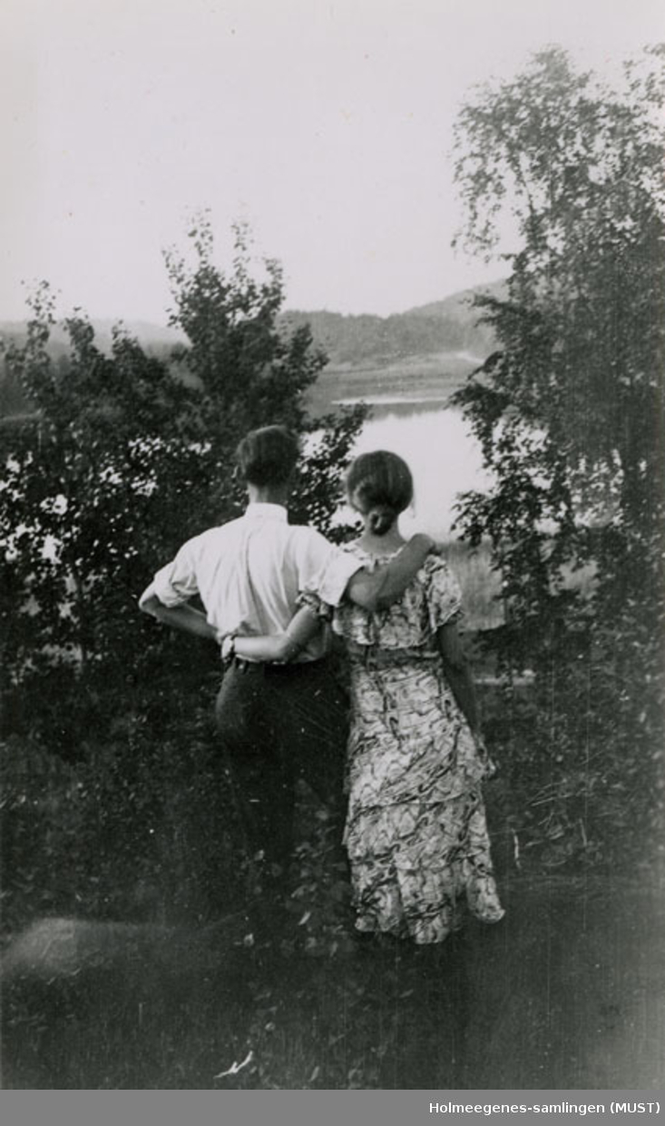 En kvinne og en mann med ryggene til fotografen står og holder om hverandre og ser ut over skog og vann i bakgrunnen av motivet. Se også ST.K.HE 2007-011-0127 til -0133.