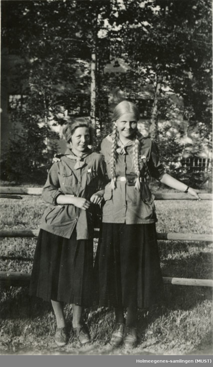 To speidere (jenter) i speiderdrakt oppstilt foran kamera. Et skogholt i bakgrunnen. Håndskrevet på baksiden av bildet: "Solveig Skavang og en venninde".