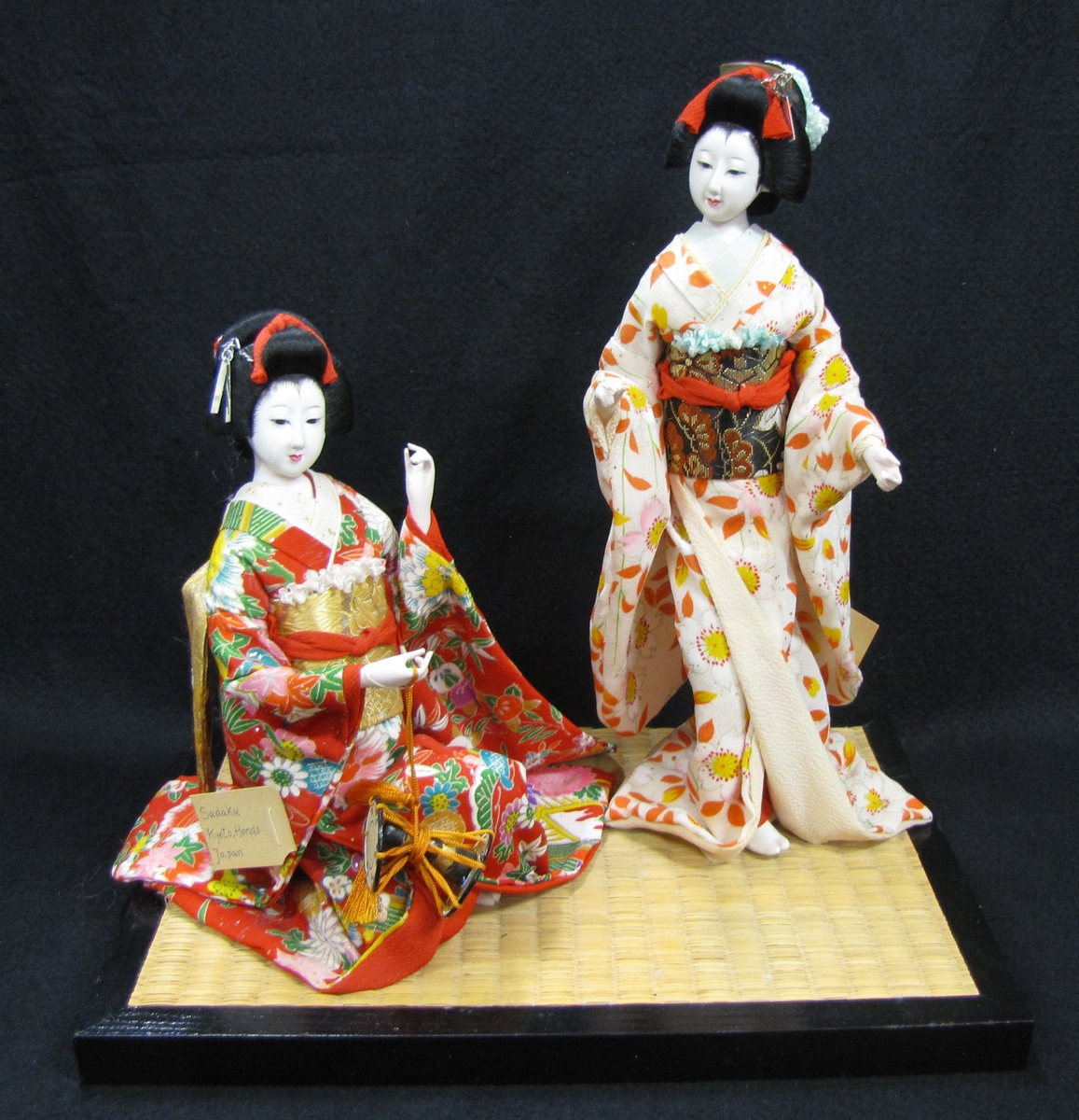 Tillhör Vera Hanssons docksamling.

Köpt 1959. Sadaku och Kikue. Kyoto, Hondo. Japan.
På en träplatta klädd med rismatta sitter en japanska i röda

kläder, en annan står upp och har solfjäder i handen.

X  Japanese Doll by Ijuin Handicraft. Ass´n.