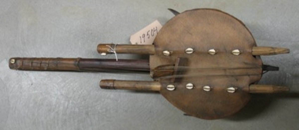 Afrikanskt stränginstrument köpt av givaren i södra Senegal 1982.
Man spelar med tummarna med instrumentet i knät, vänt från sig.
Prytt med Kaurisnäckor.