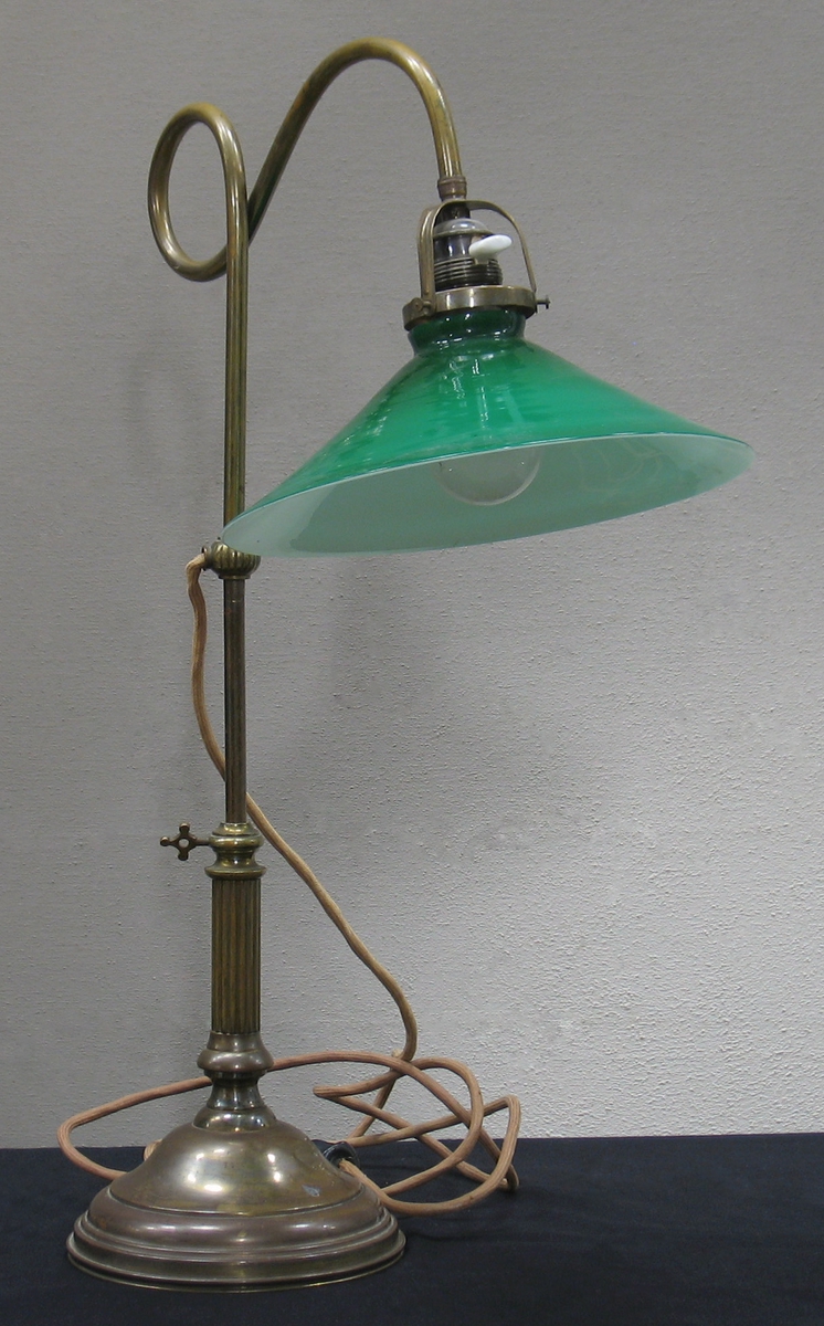 Skrivbordslampa med fot i mässing och grön glaskupa. Både fot och ställning är fint dekorerade och från mitten av ställningen löper en sladd som avslutas med stickpropp. En glödlampa är fäst innanför kupan.