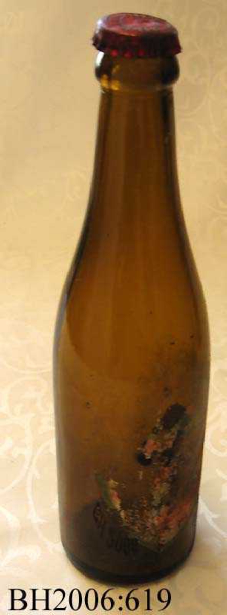 Brusflaske av brunt glass med rester av en etikett. Flasken har en orginal rød kork.