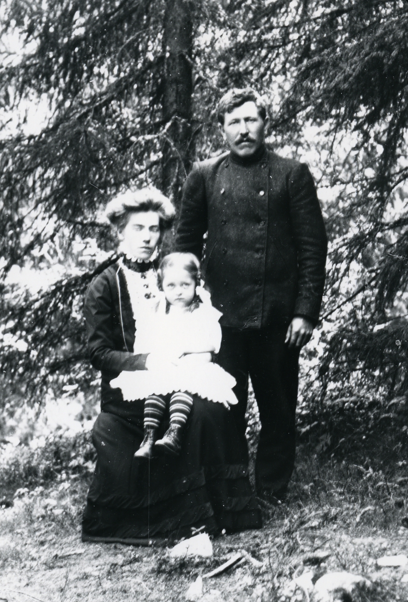 Kvinne på stol, med liten jente på fanget, og mann stående ved siden av. Skog i bakgrunnen