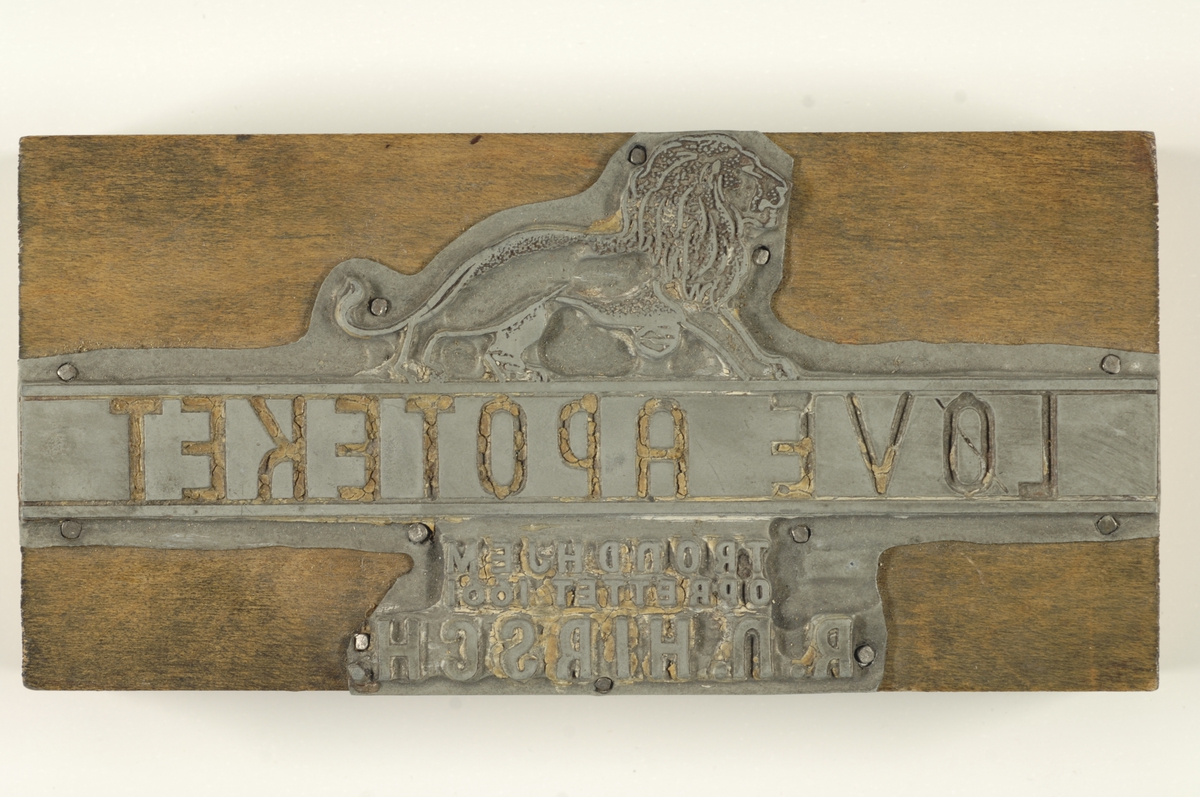 Rektangulær trekloss påstiftet en metallplate med inngravert tekst og et løvesymbol.